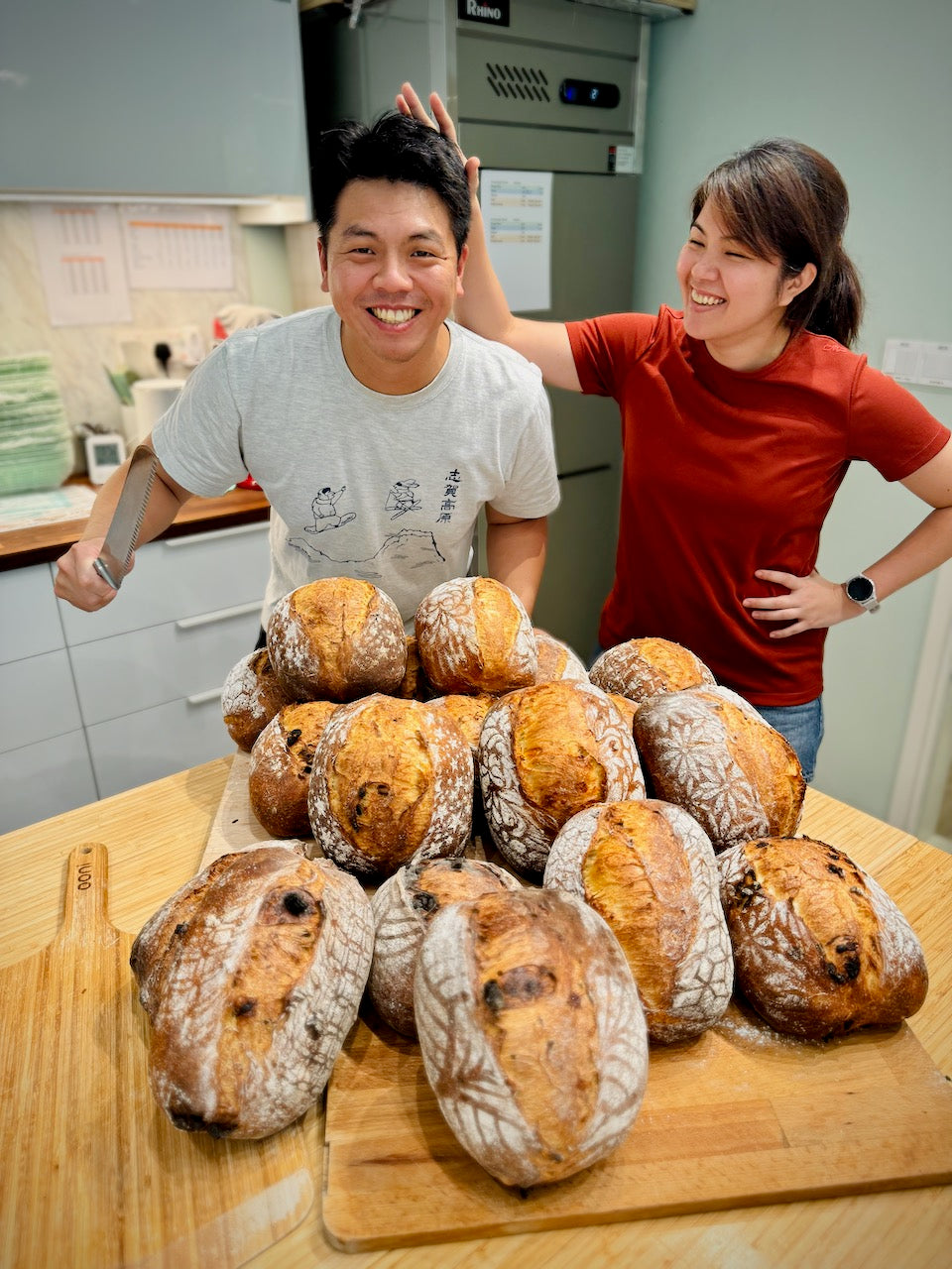 Two people standing behind a wooden cutting board with a pile of bread loaves in a kitchen.